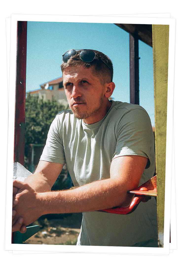 Florent Osmani blickt ernst in die Ferne. Er trägt eine Sonnenbrille auf dem Kopf. Er befindet sich draußen vor einem Haus. 