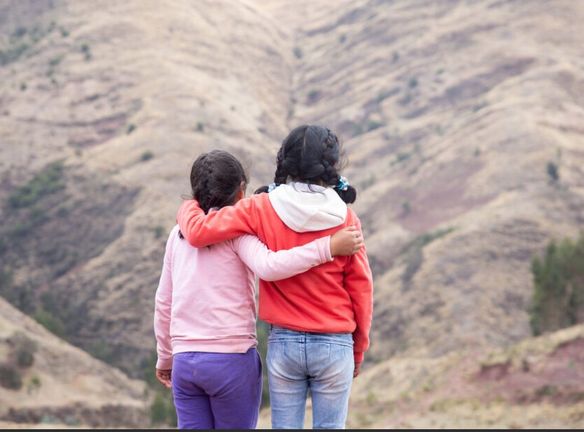 Zwei Mädchen mit Zöpfen stehen, einander umarmend und die Rücken der Kamera zugewandt, vor einer kargen Berglandschaft.