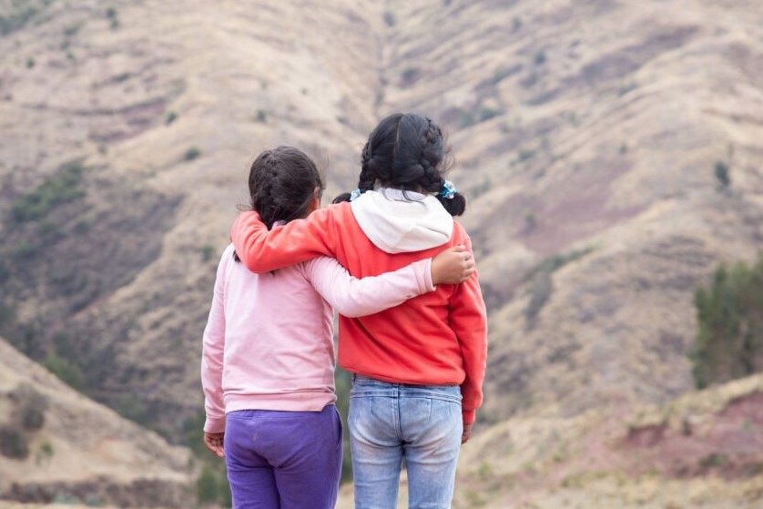 Zwei Mädchen mit Zöpfen stehen, einander umarmend und die Rücken der Kamera zugewandt, vor einer kargen Berglandschaft.