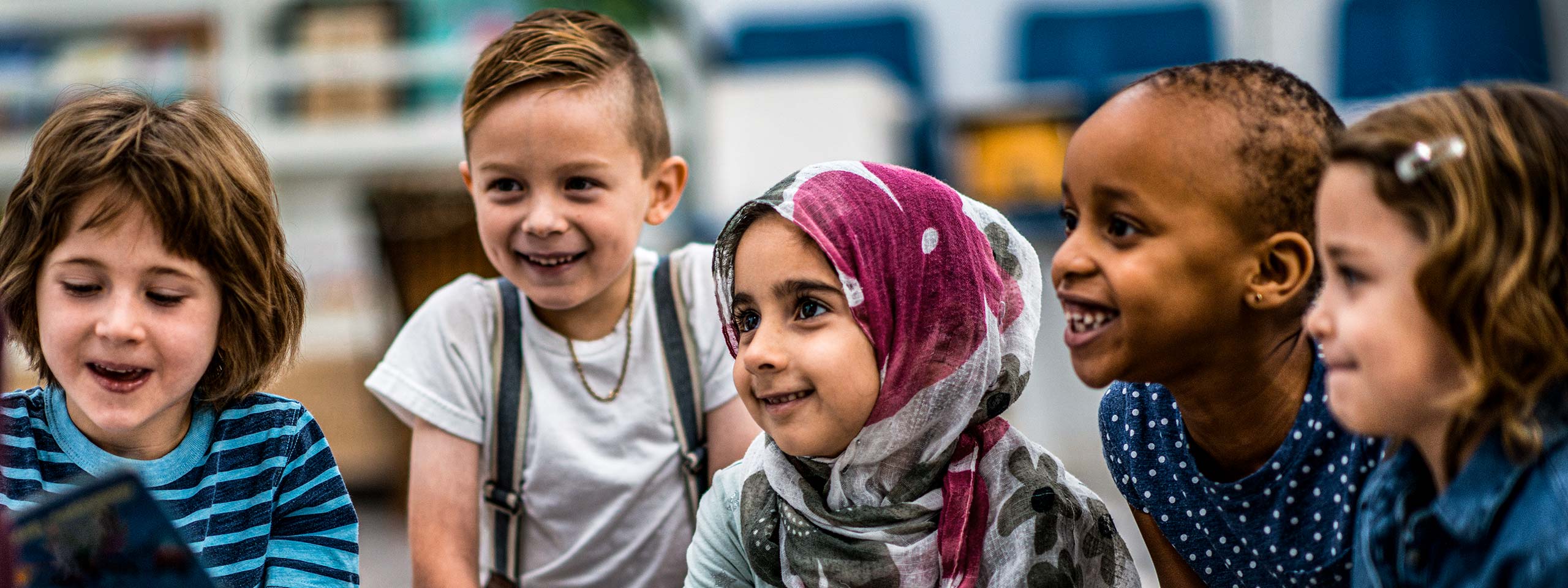 Fünf Kinder unterschiedlicher Hautfarben und Ethnien sitzen zusammen und hören zu, sie lachen. 