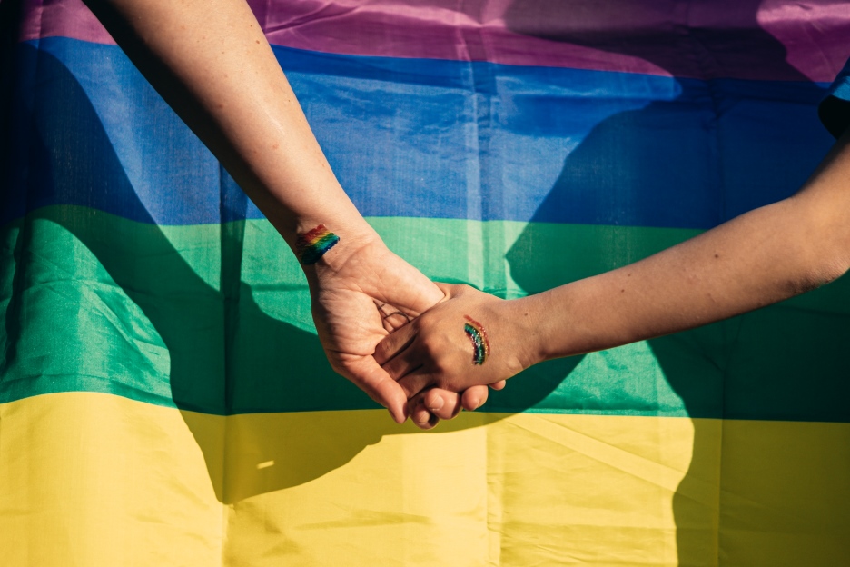 Zwei Kinderhände, die einander umfassen, im Hintergrund ist eine Regenbogenflagge zu sehen. Foto: iStock/Amparo Garcia