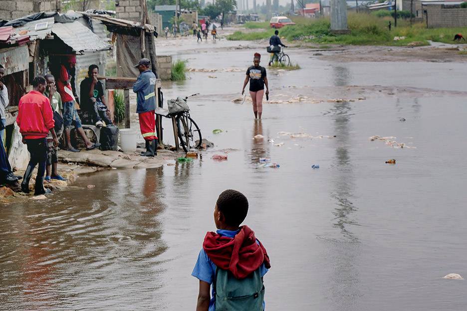 Menschen in überfluteten Gebieten Ende 2025, die durch das Hochwasser gehen oder vor Wellblechhütten stehen.