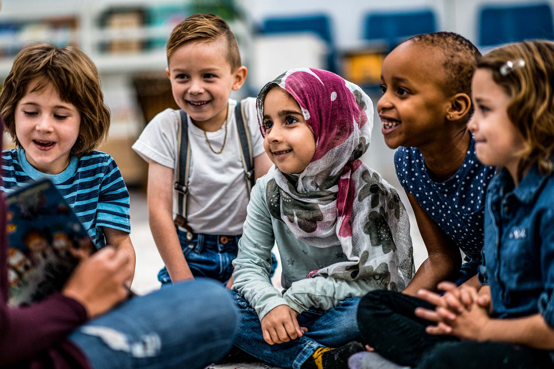 Fünf Kinder verschiedener Hautfarben und Ethnien sitzen zusammen und hören zu, sie lachen.