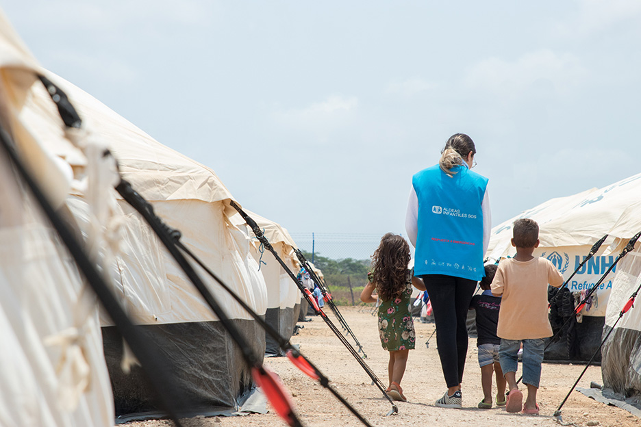 Sozialarbeiterin nimmt zwei Kinder an der Hand in einem Flüchtlingslager