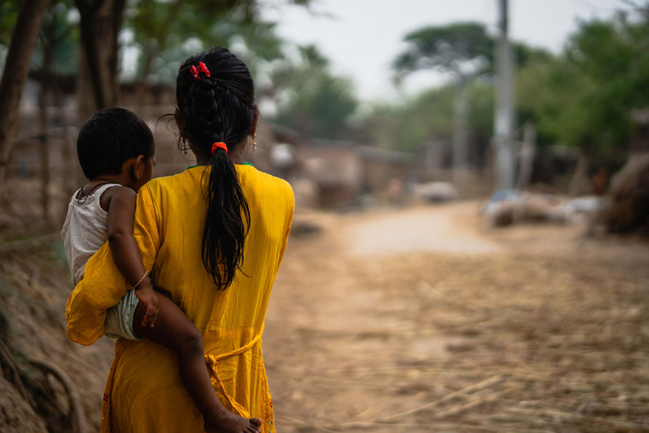 Eine Frau in gelbem Kleid trägt ein kleines Kind auf dem Arm und geht über einen unbefestigten Weg durch ein Dorf in Bangladesch
