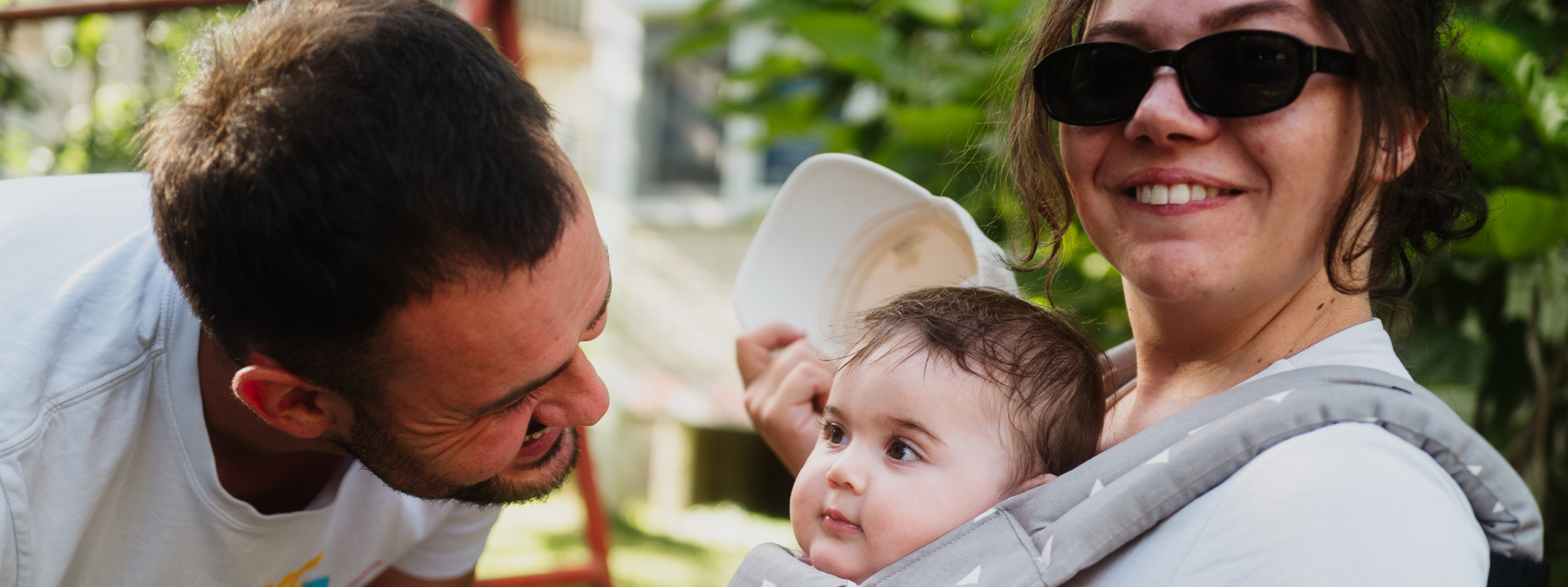 Junger Vater lächelt seine kleine Tochter an. Seine Frau trägt das Mädchen vor sich in einer Babytrage.