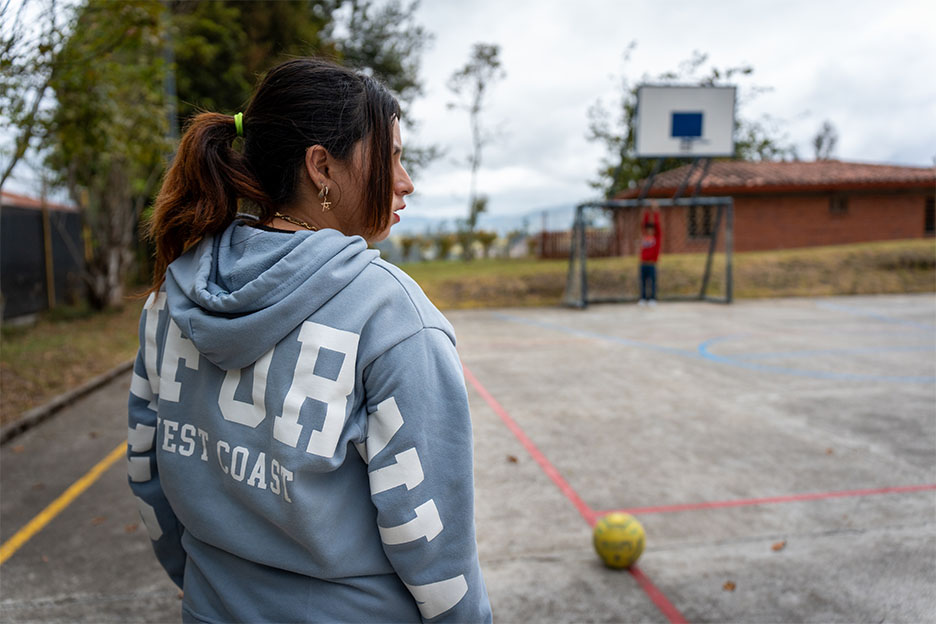 Maryan steht auf einem Sportplatz, vor ihr liegt ein Ball im Hintergrund ist ein Fußballtor, in dem ein Kind steht.