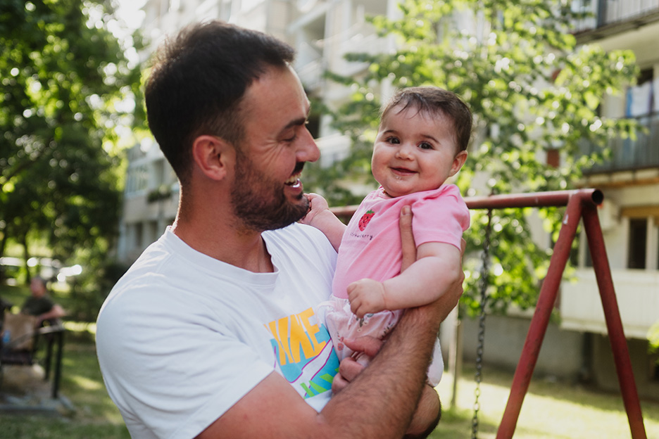 Junger Vater aus Nordmazedonien hält seine kleine Tochter im Arm. Beide lachen fröhlich.