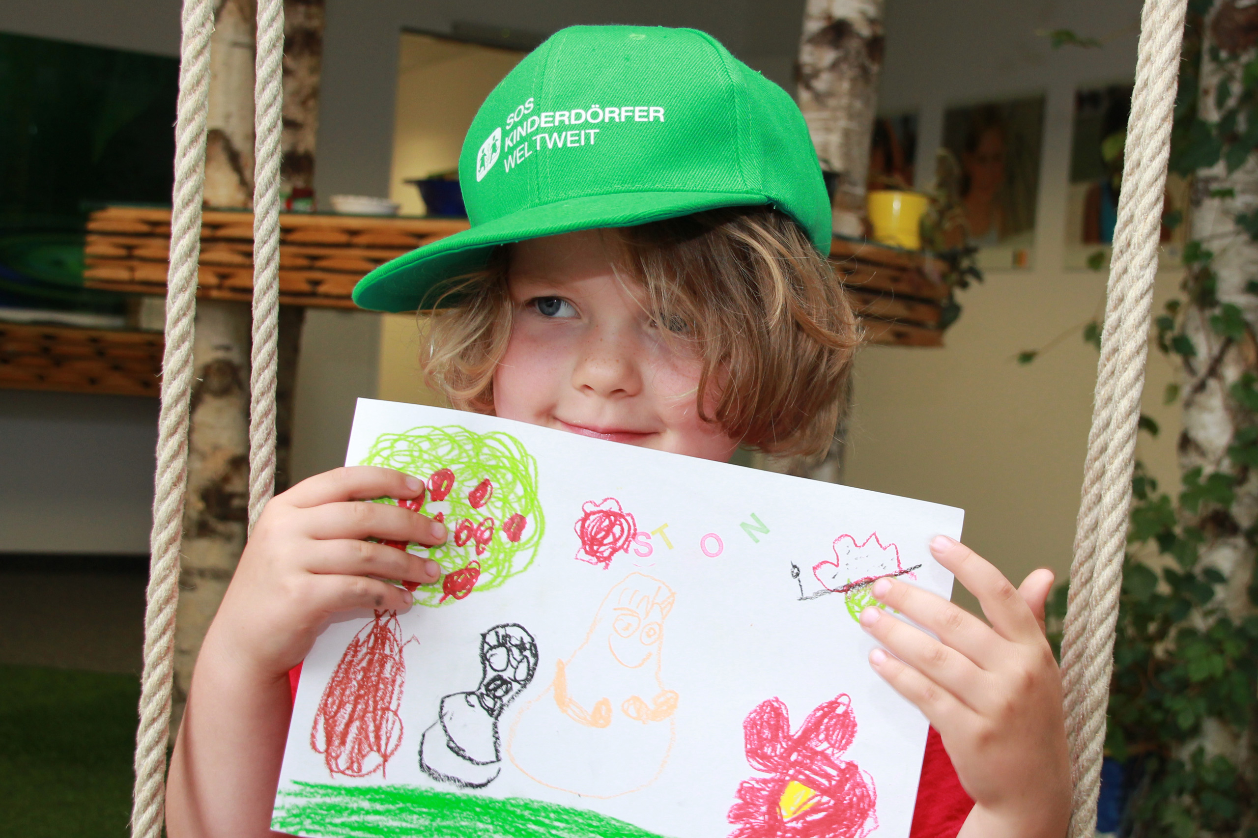 Kind mit grüner Mütze der SOS-Kinderdörfer weltweit präsentiert selbst gemaltes Bild.