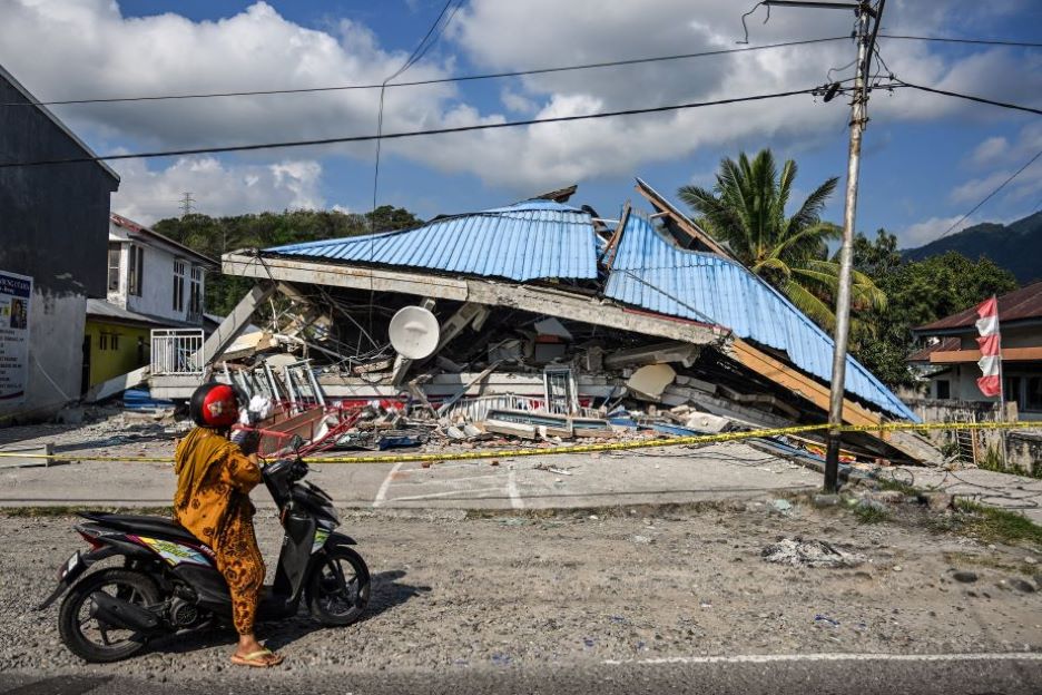 Ein eingestürztes Gebäude nach dem Erdbeben in Indonesien. Im Vordergrund steht eine Person mit einem Motorroller.