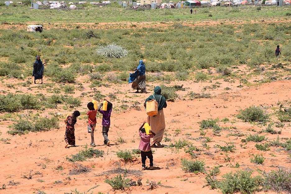 Mehrere Kinder und Frauen laufen über ein karges Feld, fast alle tragen Wasserkanister auf ihren Schultern.