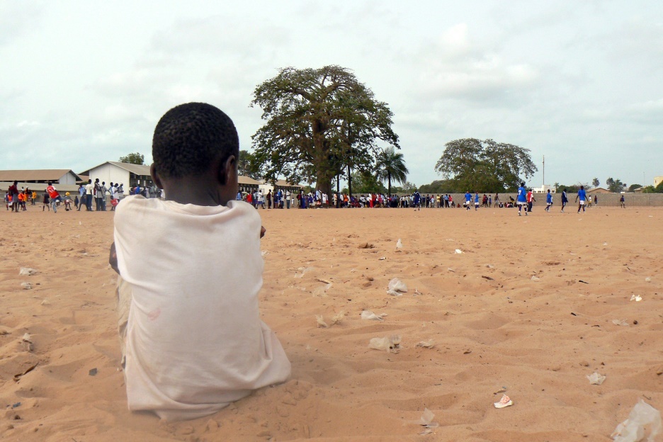 Im Vordergrund sitzt ein kleiner Junge im Sand, der anderen, von ihm weit entfernten Kindern beim Fußball spielen zusieht.