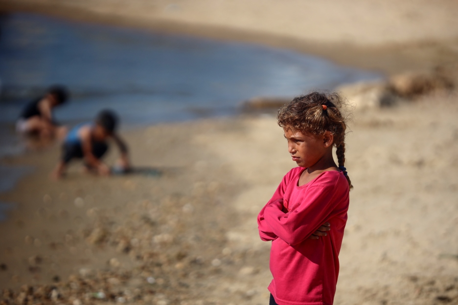 Ein junges Mädchen steht mit verschränkten Armen am Strand von Gaza, während im Hintergrund andere Kinder spielen.