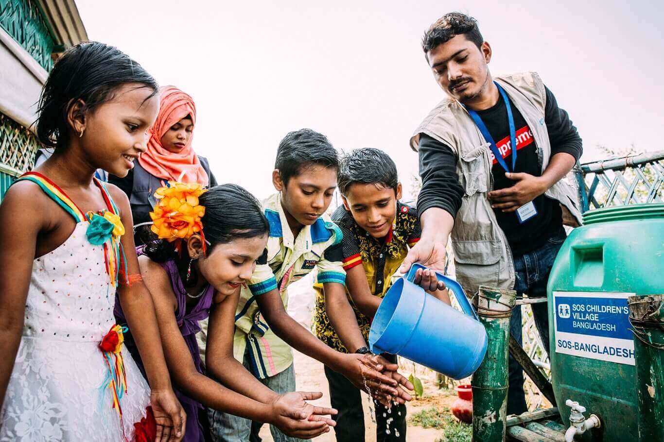 Kinder in Bangladesch waschen sich die Hände mit Wasser, das aus der Gießkanne eines SOS-Mitarbeiters fließt.