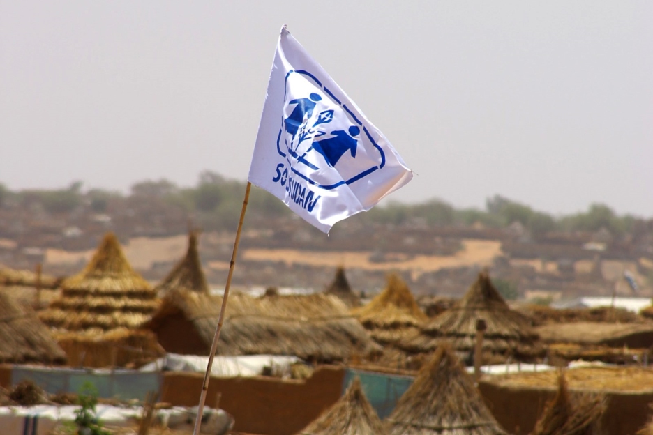Eine blau-weiße Fahne mit dem Emblem der SOS-Kinderdörfer Sudan weht über Strohdächern.  
