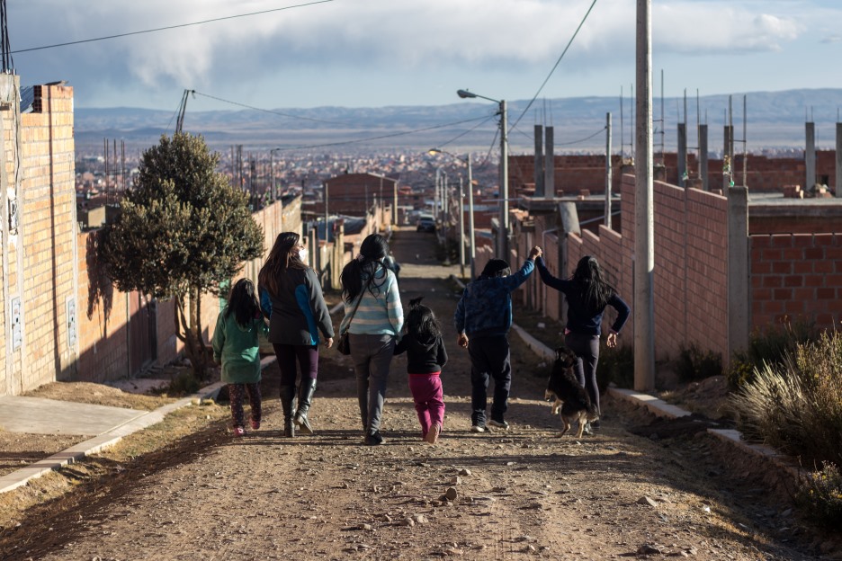 Sechs Mädchen laufen Hand in Hand, mit dem Rücken zur Kamera eine Straße entlang. Im Hintergrund sind Häuserdächer einer bolivianischen Stadt zu sehen.