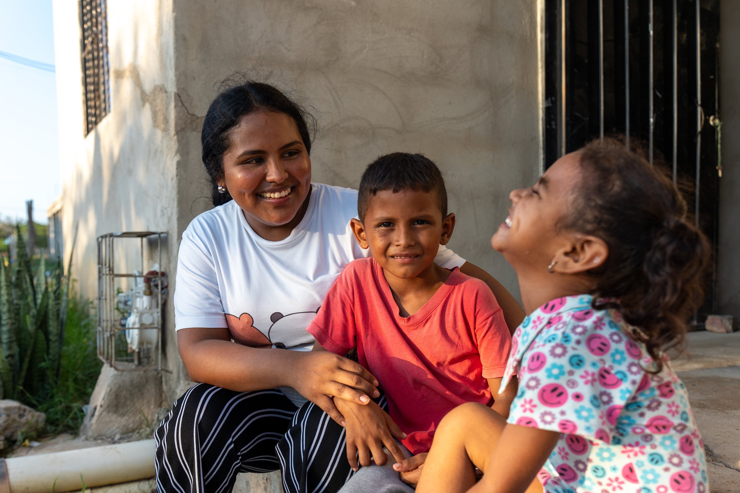 Eine junge Frau sitzt draußen neben zwei kleinen Kindern. Sie lächelt die Kinder an.