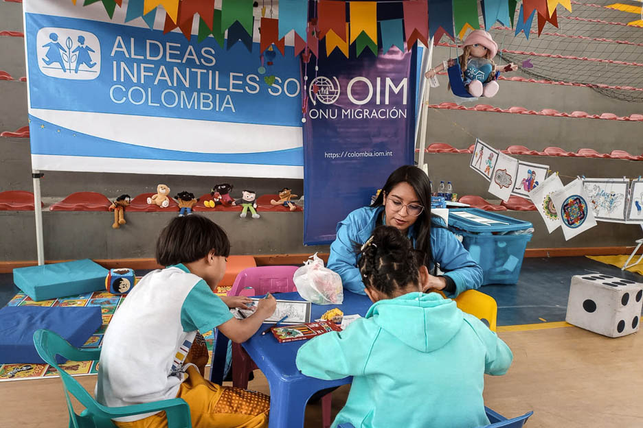 Kinder malen, Mitarbeiterin der SOS-Kinderdörfer sitzt bei ihnen. Foto: Diana Carolina Ruiz