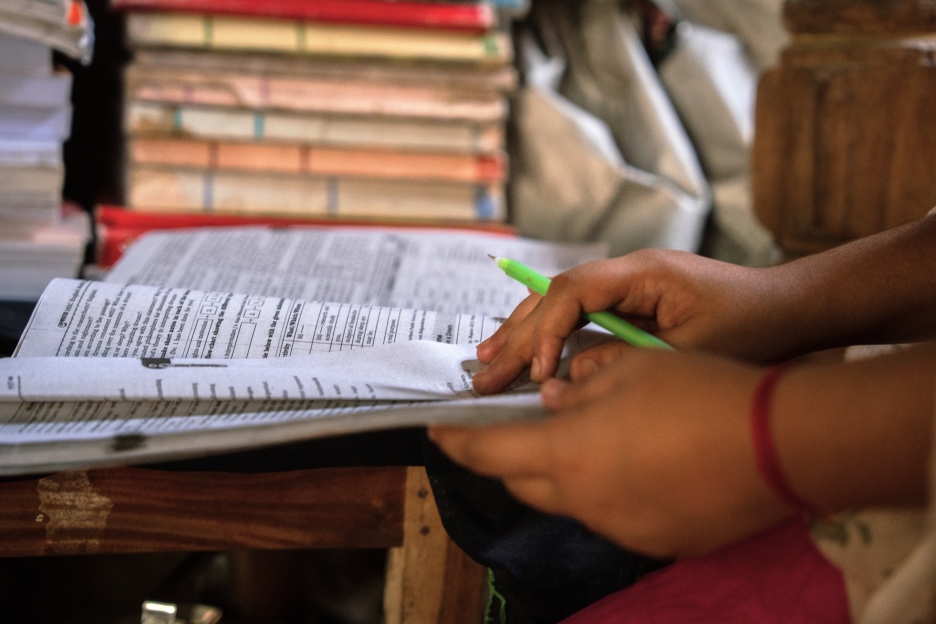 Kinderhände, die einen Stift und einen Fragebogen halten, im Hintergrund ist ein Bücherstapel zu sehen.