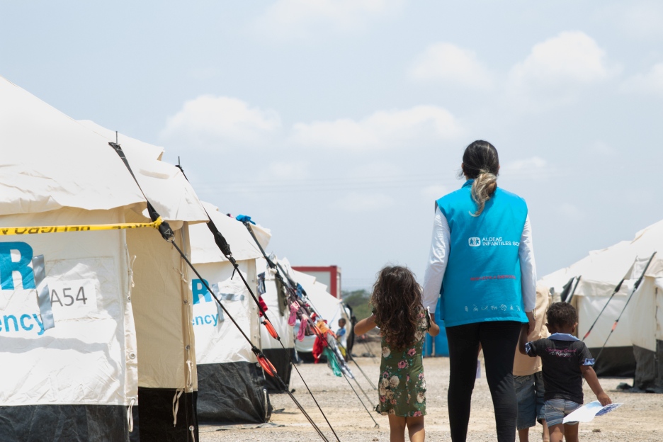 Eine humanitäre Helferin hält ein kleines Mädchen an der Hand, die beiden laufen zusammen durch ein Geflüchtetencamp der SOS-Kinderdörfer.
