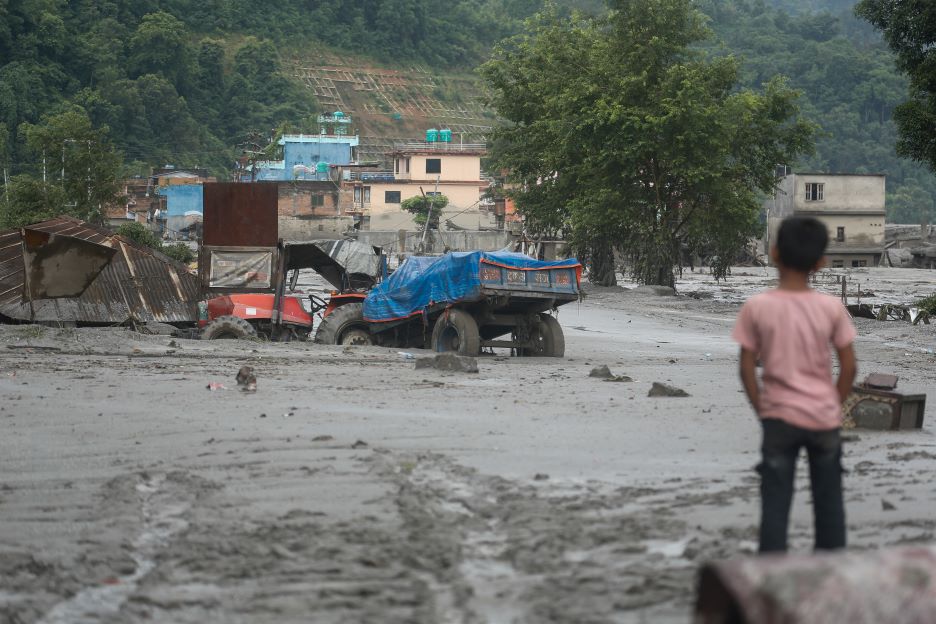 Ein Kind steht vor einer von Schlamm und Geröll bedeckten Straße nach der Sturzflut in Nepal. Im Hintergrund sind beschädigte Gebäude und Fahrzeuge zu sehen.
