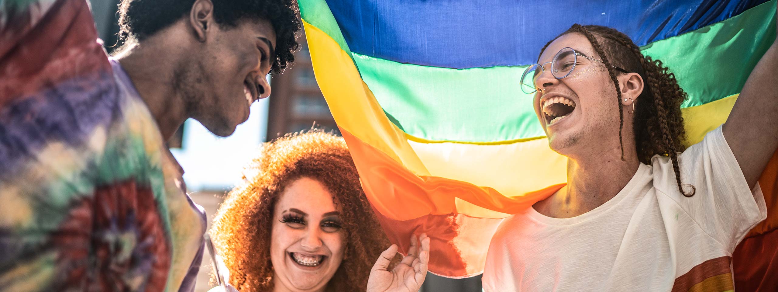 Drei junge Menschen tragen eine Regenbogenflagge und lachen ausgelassen.