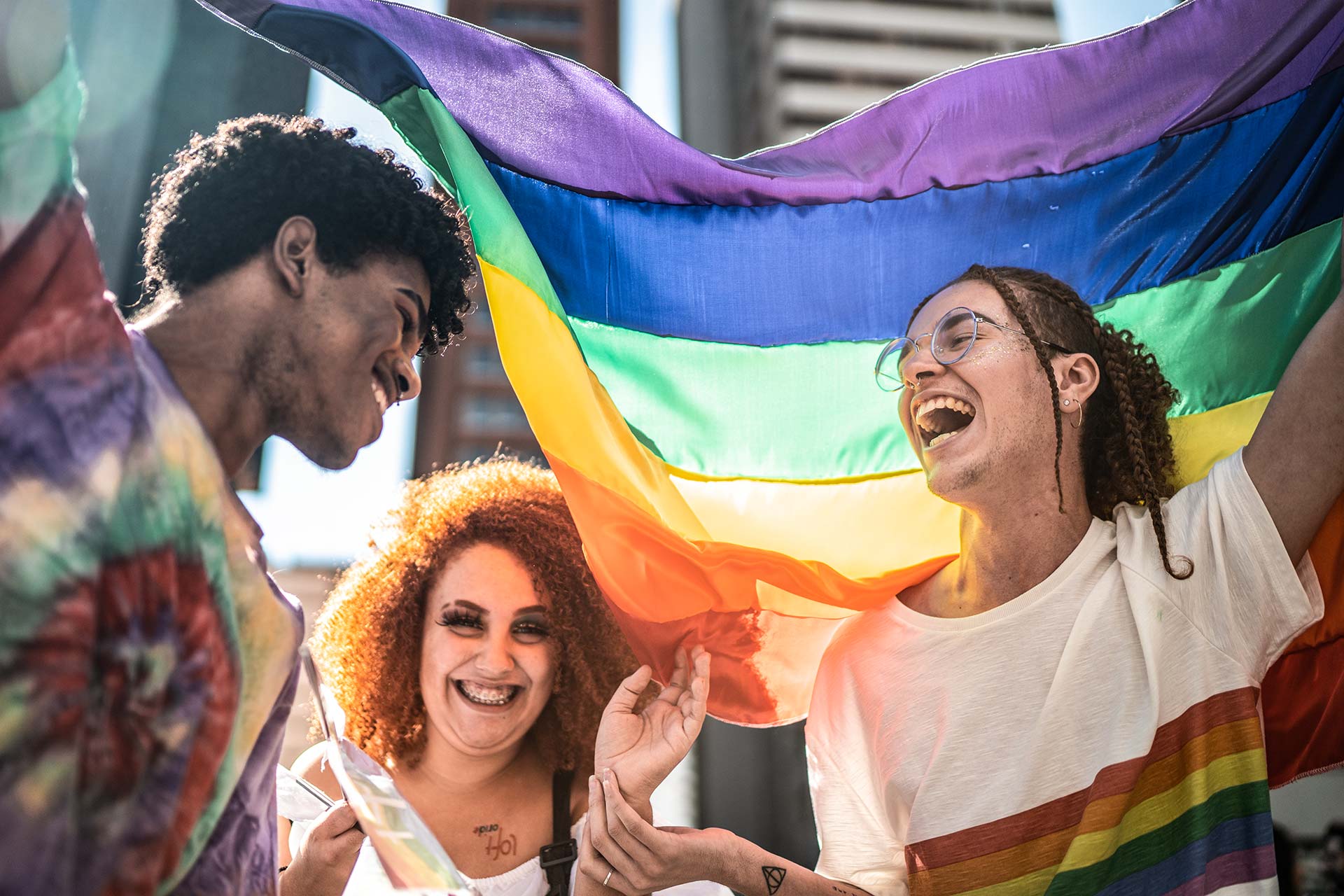 Drei junge Menschen tragen eine Regenbogenflagge, lachen ausgelassen und setzen sich für die LGBTIQ-Community ein.