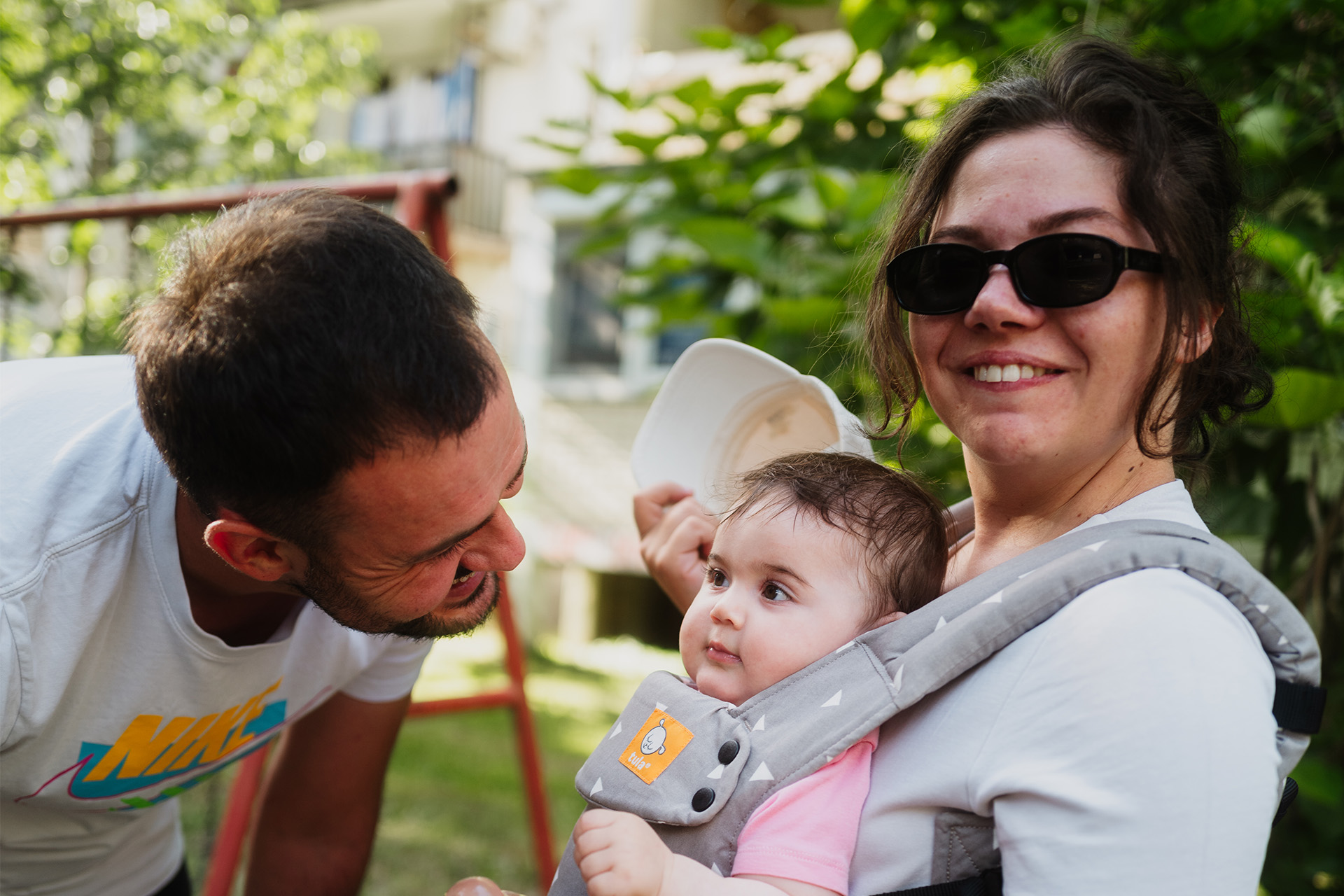 Junger Vater lächelt seine kleine Tochter an. Seine Frau trägt das Mädchen vor sich in einer Babytrage.