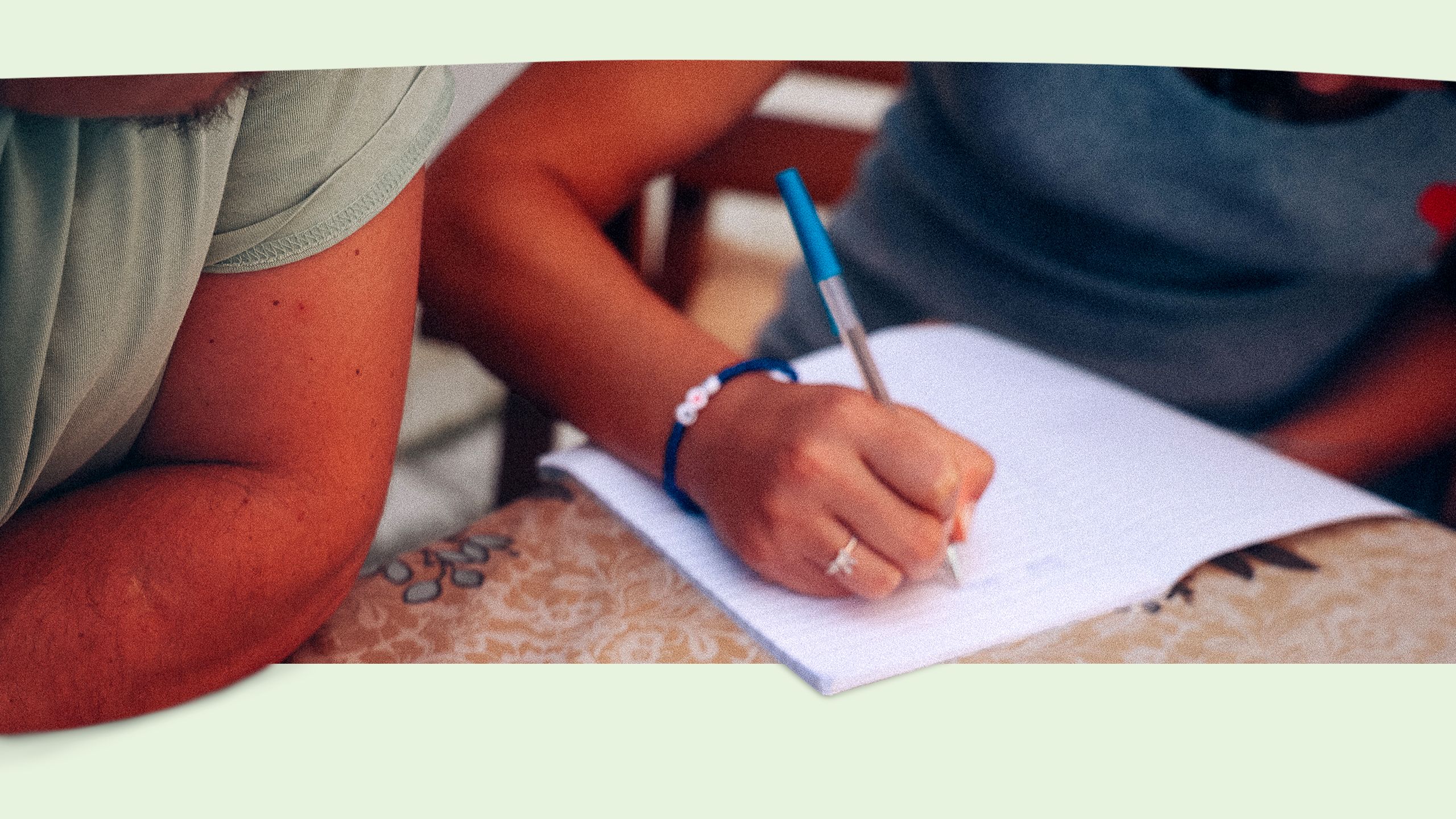 In Großaufnahme ist die Hand eines Mädchens zu sehen, das in ein Schulheft schreibt. Links im Bild ist der Arm eines Mitarbeiters des SOS-Kinderdörfer zu erkennen, der das Mädchen beim Lernen unterstützt. Beide Gesichter sind nicht sichtbar.